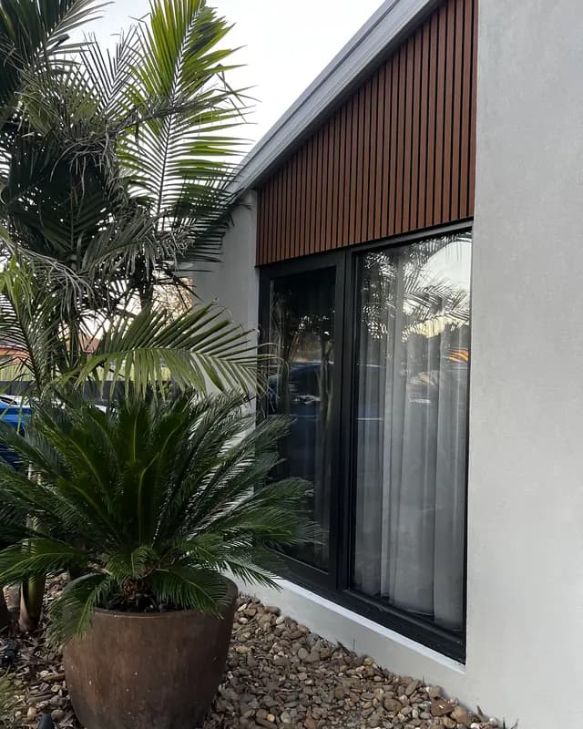 Two tall black aluminium windows in a pale rendered wall under vertical timber battens, with palms in the garden bed